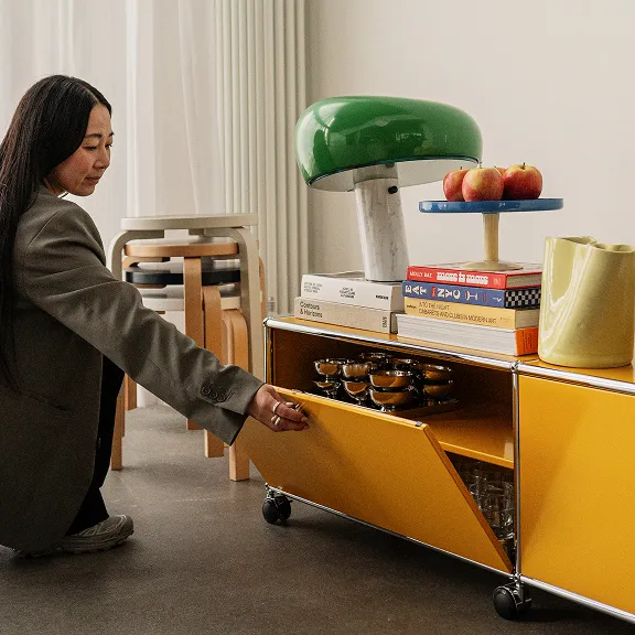 Frau in grauem Blazer öffnet Schublade eines gelben USM Haller-Regals mit Büchern, Lampe und Äpfeln in einem modernen Raum.